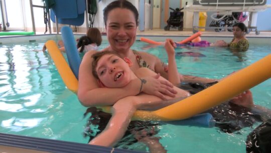 Photo of a boy with a neurodisability having hydrotherapy. He is in the pool with a specialist learning support assistant using a PODD communication book.