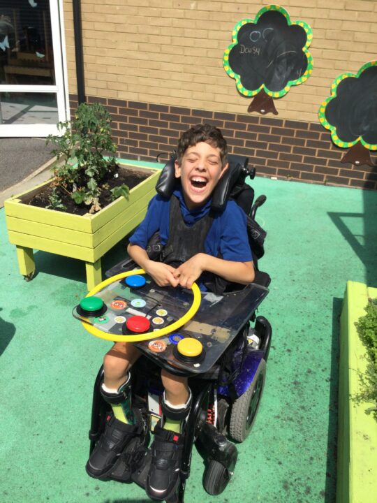 Photo of a boy with a neurodisability outside at Pace school. He is in his powered wheelchair which has 4 coloured switches on the tray, he is holding a hoop and laughing.