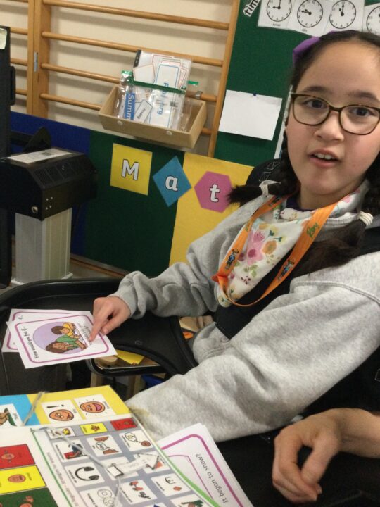 Photo of a girl with a neurodisability communicating with her teacher using a PODD communication book at Pace school.