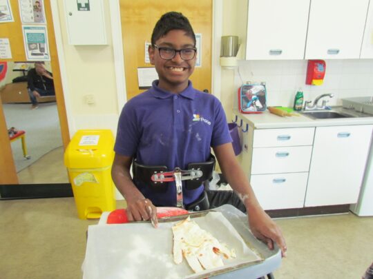 Photo of a boy with a neurodisability cooking in a daily living skills lesson at Pace school. He is in a standing frame and smiling.