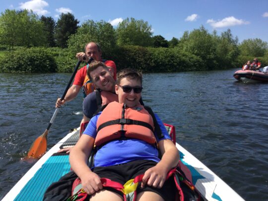 Photo of a boy with a neurodisability on a paddle board with a staff member and an instructor rowing them on a lake.