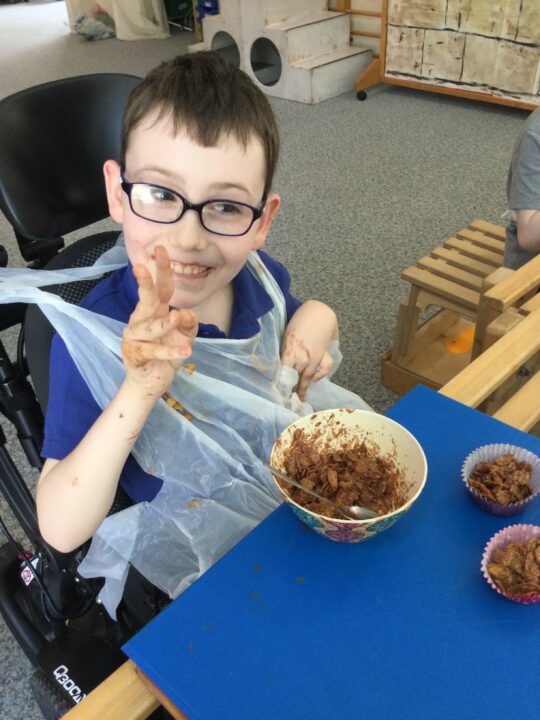 Photo of a boy with a neurodisability smiling while making chocolate cornflake cakes in a daily living skills lesson at Pace school.