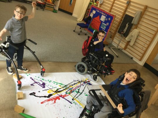 Photo of 3 boys with a neurodisability in an art lesson at Pace school. They have a canvas on the floors and creating a picture with lots of brightly coloured paints. All of the boys are laughing and smiling.