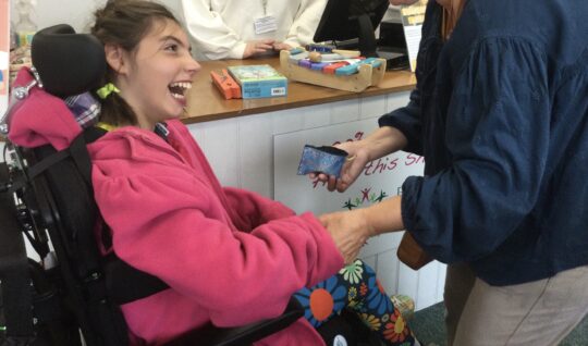 Photo of a girl with a neurodisability in a wheelchair with a Conductor. They are at the till in a charity shop counting money from a purse.
