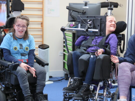 Photo of 2 girls with neurodisabilities in their class at Pace school. They are taking part in a debate and both girls are smiling. One of the girls is using an EyeGaze computer to communicate.