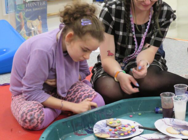 A Pace student dressed in purple looks at the biscuits she has decorated with purple icing and Smarties. The Learning Support Assistant watches her with their face just out of shot.