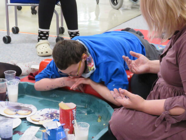 A Pace student lies on his stomach on a support ramp, looking at his plate of decorated biscuits. To the right, the teacher is partially off camera but her hands are covered in purple icing.