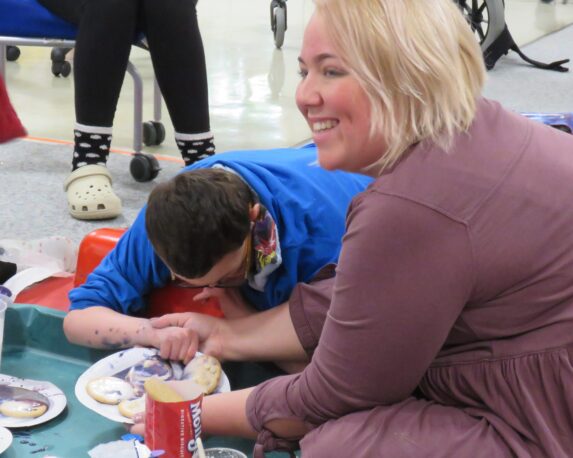 A pace student is lying on his stomach on a support ramp while holding the paper plate with his decorated biscuits. The class teacher is smiling while looking to the side, holding his hand.