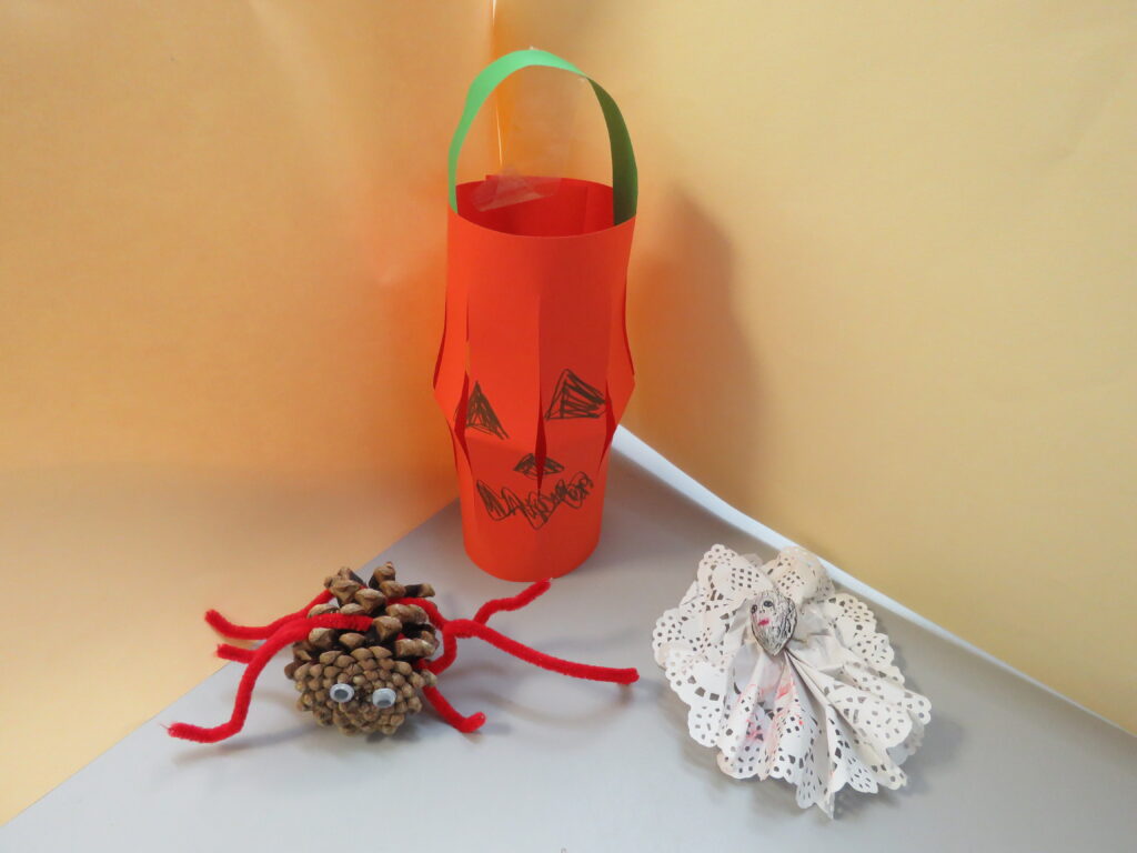 Three hand made Halloween decorations. A spider made from a pinecone with red pipe cleaner legs and two google eyes. A paper lantern with a spooky pumpkin face.  A ghost made with decorative paper.