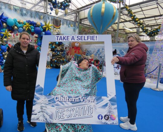 A person in a wheelchair wrapped in a patterned blanket poses behind a large “TAKE A SELFIE! Chiltern View ICE SKATING” frame, held by two adults in a winter-themed, holiday-decorated venue.
